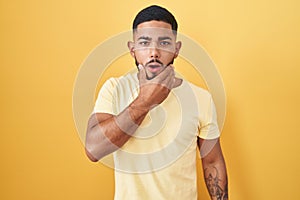 Young hispanic man standing over yellow background looking fascinated with disbelief, surprise and amazed expression with hands on