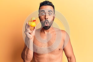 Young hispanic man standing shirtless holding duck toy scared and amazed with open mouth for surprise, disbelief face