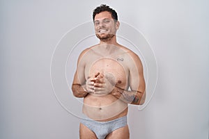 Young hispanic man standing shirtless wearing underware hands together and fingers crossed smiling relaxed and cheerful