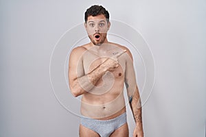 Young hispanic man standing shirtless wearing underware surprised pointing with finger to the side, open mouth amazed expression