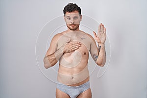 Young hispanic man standing shirtless wearing underware swearing with hand on chest and open palm, making a loyalty promise oath