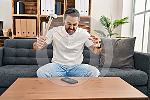 Young hispanic man stressed breaking smartphone with hammer at home