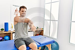 Young hispanic man stretching arm sitting on massage board at clinic