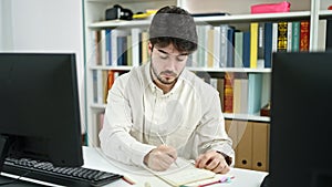 Young hispanic man student using computer writing notes at library university