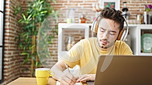 Young hispanic man student using laptop and headphones taking notes at dinning room