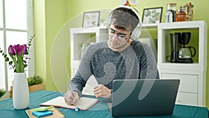 Young hispanic man student using laptop taking notes wearing headphones at dinning room