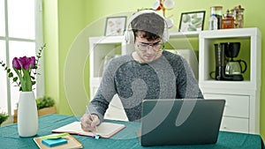 Young hispanic man student using laptop taking notes wearing headphones at dinning room