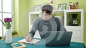Young hispanic man student using laptop taking notes wearing headphones at dinning room