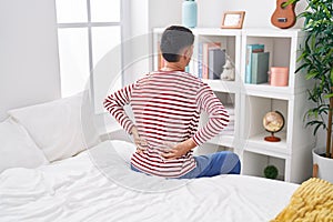 Young hispanic man suffering for back injury sitting on bed at bedroom