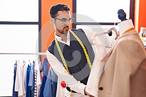 Young hispanic man tailor holding t shirt standing by manikin at atelier