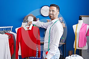 Young hispanic man tailor smiling confident holding clothes on rack at sewing studio