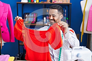 Young hispanic man tailor smiling confident holding clothes at sewing studio