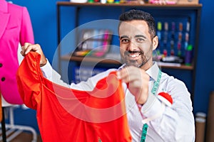 Young hispanic man tailor smiling confident holding clothes at sewing studio