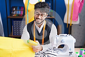 Young hispanic man tailor smiling confident holding cloths and thread at sewing studio