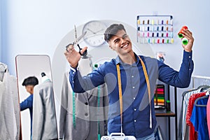 Young hispanic man tailor smiling confident holding scissors and thread at clothing factory