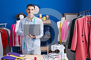 Young hispanic man tailor smiling confident using laptop at sewing studio