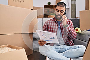 Young hispanic man talking on smartphone reading document at new home