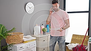 Young hispanic man talking on smartphone reading washing machine instructions at laundry room