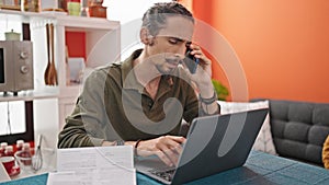 Young hispanic man talking on smartphone using laptop at dinning room