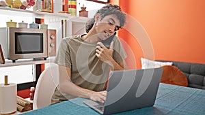 Young hispanic man talking on smartphone using laptop at dinning room
