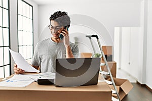 Young hispanic man talking on the smartphone using laptop at new home