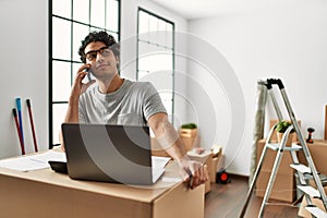 Young hispanic man talking on the smartphone using laptop at new home