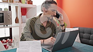 Young hispanic man talking on smartphone using laptop reading document at dinning room