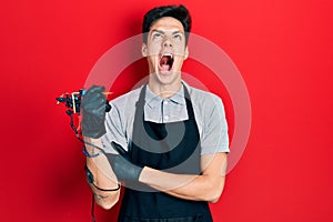 Young hispanic man tattoo artist wearing professional uniform and gloves holding tattooer machine annoyed and frustrated shouting