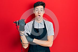 Young hispanic man tattoo artist wearing professional uniform and gloves holding tattooer machine covering mouth with hand,