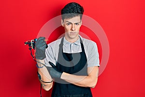 Young hispanic man tattoo artist wearing professional uniform and gloves holding tattooer machine smiling happy and positive,