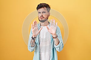 Young hispanic man with tattoos standing over yellow background moving away hands palms showing refusal and denial with afraid and
