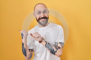 Young hispanic man with tattoos standing over yellow background pointing to the back behind with hand and thumbs up, smiling