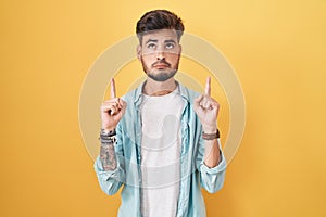 Young hispanic man with tattoos standing over yellow background pointing up looking sad and upset, indicating direction with