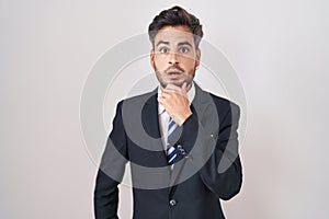 Young hispanic man with tattoos wearing business suit and tie looking fascinated with disbelief, surprise and amazed expression