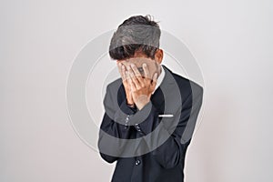 Young hispanic man with tattoos wearing business suit and tie with sad expression covering face with hands while crying