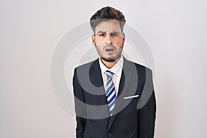 Young hispanic man with tattoos wearing business suit and tie in shock face, looking skeptical and sarcastic, surprised with open