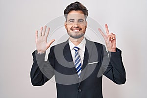 Young hispanic man with tattoos wearing business suit and tie showing and pointing up with fingers number seven while smiling