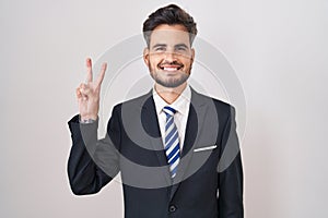 Young hispanic man with tattoos wearing business suit and tie showing and pointing up with fingers number two while smiling