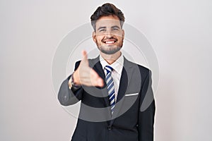 Young hispanic man with tattoos wearing business suit and tie smiling friendly offering handshake as greeting and welcoming