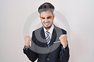 Young hispanic man with tattoos wearing business suit and tie very happy and excited doing winner gesture with arms raised,