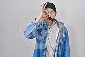 Young hispanic man with tattoos wearing wool cap doing ok gesture shocked with surprised face, eye looking through fingers