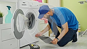 Young hispanic man technician repairing washing machine reading instructions at laundry room