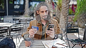 Young hispanic man tourist shopping with smartphone and credit card sitting on table at coffee shop terrace