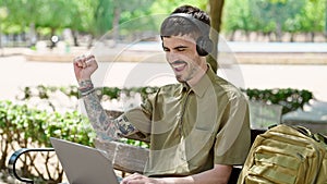 Young hispanic man tourist using laptop and headphones celebrating at park