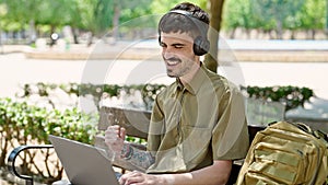Young hispanic man tourist using laptop and headphones celebrating at park