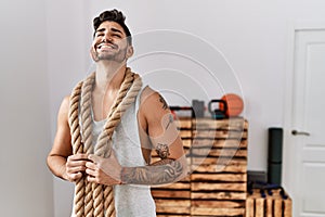 Young hispanic man training with battle rope at the gym smiling and laughing hard out loud because funny crazy joke