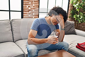 Young hispanic man unwell drinking water sitting on sofa at home