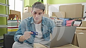 Young hispanic man using laptop and credit card sitting on sofa at new home