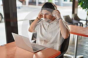 Young hispanic man using laptop and headphones at coffee shop terrace