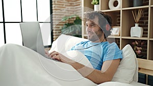 Young hispanic man using laptop and headphones lying on bed at bedroom